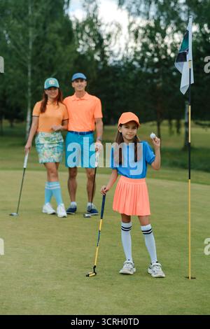 Une famille passe sa journée à jouer au mini-golf, avec la fille prête à putt et ses parents souriant en arrière-plan. Ils portent des tenues lumineuses, réf Banque D'Images