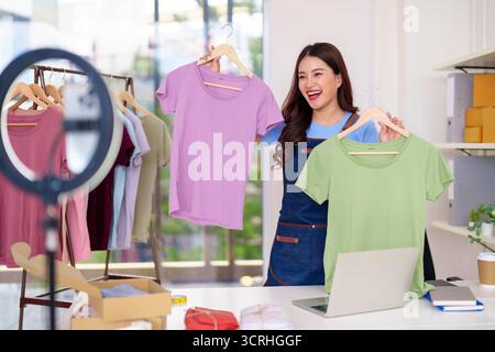 Heureuse femme adulte asiatique présentant des t-shirts colorés dans sa configuration de home studio pour un flux en direct vendant des vêtements en ligne via un ordinateur portable Banque D'Images