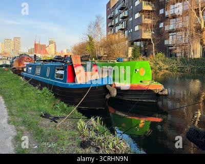 Deux bateaux étroits animés amarrés au bord d'un canal urbain, adjacents à des bâtiments modernes et à un paysage urbain, entourés de verdure et réfléchissant sur l'eau calme, Banque D'Images