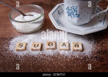 Le mot sucre avec une pincée de sucre blanc avec une tasse à thé et une soucoupe, et le sucre et la cuillère à thé dans un récipient en verre. Banque D'Images