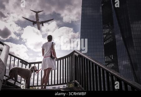 Femme avec Dog Watching Airplane volant au-dessus de City Skyscraper, scène urbaine symbolisant le voyage, la liberté et la vie moderne Banque D'Images