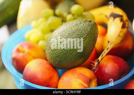 Avocat couché sur le dessus des fruits mélangés dans un bol bleu. Gros plan sur l'assortiment de fruits tropicaux. Concept de nutrition saine et de vitamines naturelles. Banque D'Images