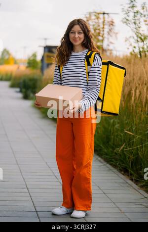 Jeune femme souriante travaillant comme coursier, portant un pantalon orange vif et une chemise rayée, portant un paquet de carton vierge et un grand sac thermique jaune, livrant de la nourriture ou des colis Banque D'Images
