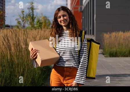 Femme livreuse tenant un colis, portant une chemise rayée et un sac à dos de livraison jaune, travaillant dans une zone urbaine, représentant le shopping en ligne et une économie de concert Banque D'Images