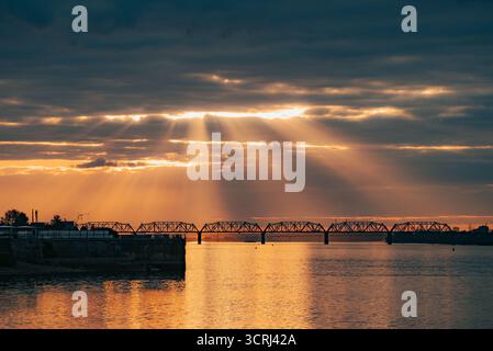 Pont enjambant une large rivière au coucher du soleil, avec un paysage nuageux sombre créant un contraste saisissant avec des rayons de soleil brillants illuminant le ciel et reflétant une lueur dorée à travers la surface de l'eau Banque D'Images