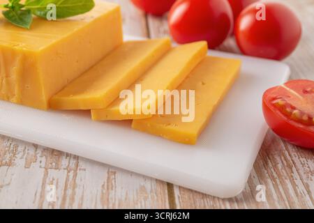 Gros plan sur les tranches de fromage Sheddar. Fromage orange fendu sur une planche à découper blanche et quelques tomates rouges sur une table en bois. Produit laitier, calcium. Banque D'Images