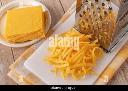 Gros plan de fromage cheddar entier et râpé. Fromage orange râpé et râpé sur une planche à découper blanche près de la pièce entière sur une assiette. Produit laitier. Banque D'Images