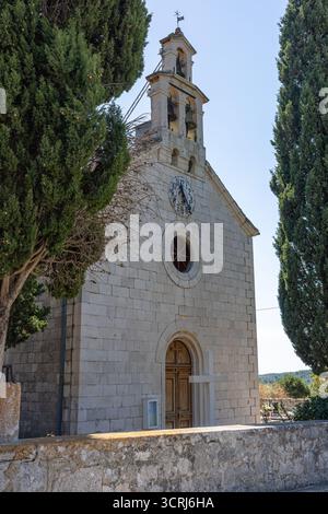 Une petite église en pierre avec un cadran d'horloge et des clochers jumeaux, encadrée par deux cyprès imposants contre un ciel bleu vif. Un R serein et historique Banque D'Images