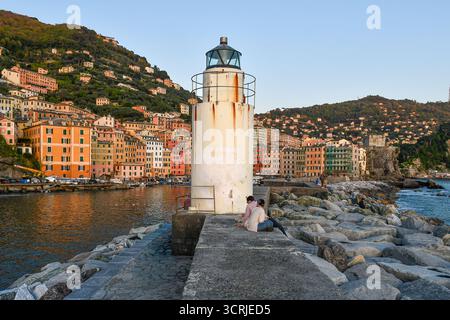 Le phare blanc sur la jetée brise-lames, avec le village de pêcheurs coloré en arrière-plan au coucher du soleil, Camogli (Gênes), Ligurie, Italie Banque D'Images