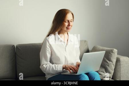 Heureuse femme mature moderne travaillant avec un ordinateur portable, fille freelance regardant l'ordinateur assis sur un canapé dans le salon à la maison Banque D'Images