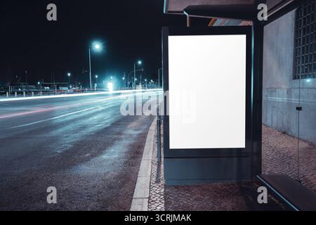 Panneau publicitaire éclairé vierge à un abri de bus à côté d'une route humide de la ville la nuit, capturé avec des traînées lumineuses de voiture à longue exposition et une rue lumineuse Banque D'Images