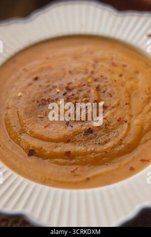 Soupe chaude et crémeuse à l'orange servie dans un bol blanc, garnie de flocons de piment rouge et de poivre noir Banque D'Images