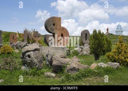 Sculptures en pierre en forme de lettres sur un pré vert, entouré de rochers et de buissons sous un ciel nuageux, monument à l'alphabet arménien, Apar Banque D'Images