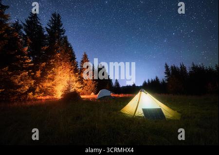 Tente jaune à côté du panneau solaire sous le ciel étoilé magnifique. À proximité, d'autres tentes et arbres sont éclairés par la lumière chaude du feu de camp, soulignant l'expérience de camping écologique et sereine. Banque D'Images