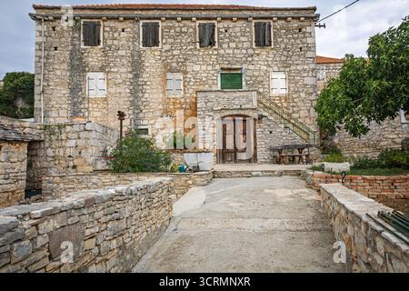 Bâtiment en pierre altérée dans une cour ensoleillée avec une porte en bois, des marches en pierre et une végétation luxuriante dans un cadre de village rustique. Banque D'Images