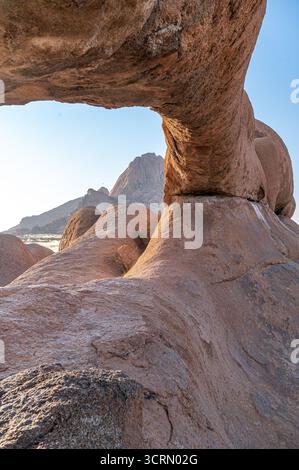 Vue grand angle à travers la spectaculaire arche rocheuse de granit à Spitzkoppe, rochers anciens encadrant des pics montagneux accidentés sous un ciel bleu vif, Namibie Banque D'Images