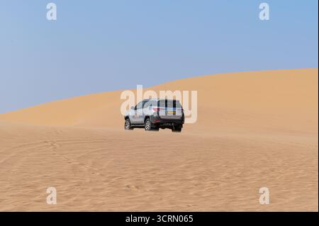 Vue arrière de Toyota Fortuner SUV blanc montant dune dorée abrupte Namib, vaste étendue désertique et fond de ciel bleu, Sandwich Harbour, Namibie. Banque D'Images
