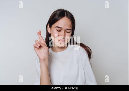 Jeune femme souriante croisant les doigts pour bonne chance dans un cadre de studio léger Banque D'Images