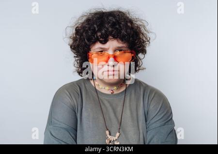 Un individu aux cheveux bouclés portant des lunettes de soleil orange vif regarde attentivement l'appareil photo tout en portant une tenue décontractée. Les accessoires colorés ajoutent a u Banque D'Images