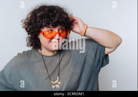 Une personne aux cheveux bouclés met en valeur son style unique en portant des lunettes de soleil orange et des accessoires colorés. Ils posent de manière ludique dans un e intérieur détendu Banque D'Images