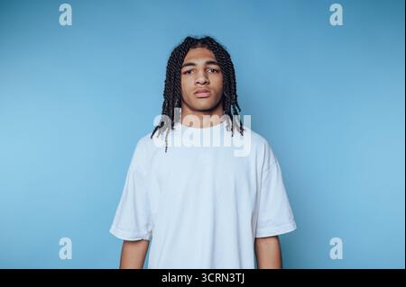 Un jeune homme se tient droit contre un fond bleu solide. Il porte une chemise blanche lâche, mettant en valeur sa coiffure tressée tout en affichant son expression neutre Banque D'Images