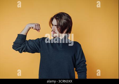 Un jeune homme exprime sa confiance en fléchissant son bras, mettant en valeur son biceps sur un fond jaune vif, vêtu d'un pull sombre décontracté, Conve Banque D'Images