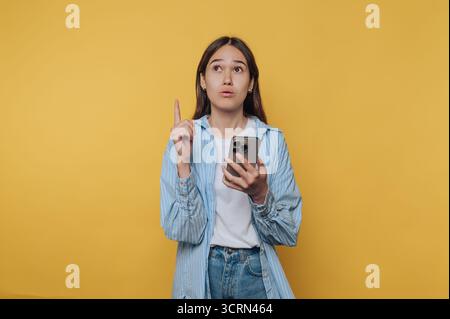 Une jeune femme tenant un smartphone semble réfléchie, levant son index comme si elle avait une idée, sur un fond jaune Uni. Banque D'Images