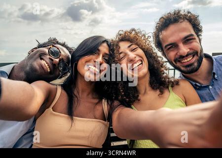 Groupe multiculturel de quatre jeunes amis prenant un selfie joyeux en plein air au bord de la mer, riant et profitant de vacances d'été ensemble. Banque D'Images