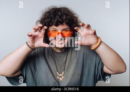 Une personne aux cheveux bouclés portant des lunettes de soleil orange pose de manière ludique avec des gestes de la main en forme de griffe, souriant largement. Ils portent un t-shirt gris, coloré b Banque D'Images