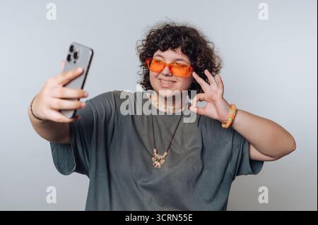 Une personne aux cheveux bouclés portant des lunettes de soleil orange prend un selfie, faisant un geste OK avec sa main. Ils portent un t-shirt gris et coloré Banque D'Images
