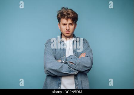 Jeune homme avec les bras croisés et une expression sceptique sur fond bleu. Banque D'Images