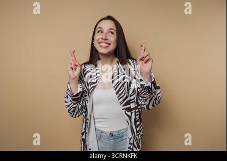 Une femme souriante avec de longs cheveux croisant les doigts, portant une veste à motif zèbre et un Jean bleu clair, debout sur un fond beige. Banque D'Images