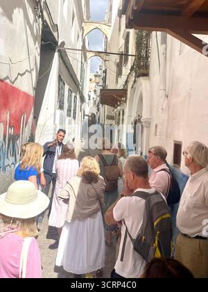 Les ruelles sont étroites et sinueuses dans les zones historiques des villes médiévales nord-africaines et arabes. Les touristes aiment marcher dans une médina ou un souk car ils offrent beaucoup de culture et d'histoire. Un jeune homme local guide un groupe de touristes anglais à travers la vieille ville de Tanger, au Maroc. Banque D'Images