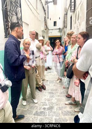 Les ruelles sont étroites et sinueuses dans les zones historiques des villes médiévales nord-africaines et arabes. Les touristes aiment marcher dans une médina ou un souk car ils offrent beaucoup de culture et d'histoire. Un jeune homme local guide un groupe de touristes anglais à travers la vieille ville de Tanger, au Maroc. Banque D'Images