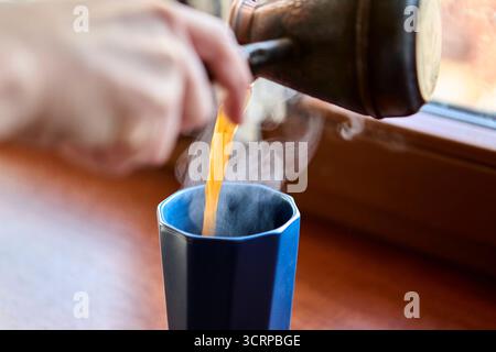 Vue rapprochée d'un café chaud fumant versé dans une grande tasse en céramique foncée placée sur le rebord de la fenêtre. La vapeur monte pour créer une ambiance maison confortable. Banque D'Images