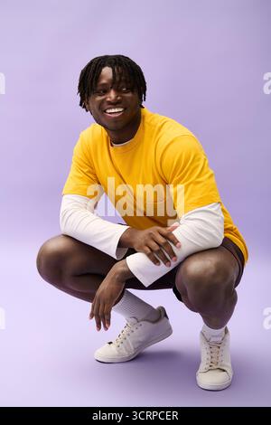 Un jeune homme portant une chemise jaune vif et un short sourit tout en accroupissant sur un fond vibrant. Banque D'Images