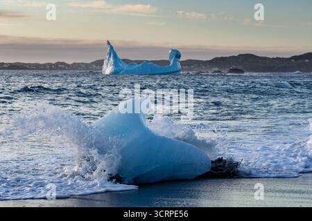 Un petit iceberg se trouve sur une plage de sable noir tandis que les vagues s'écrasent autour d'elle, avec un iceberg plus grand visible en arrière-plan, mettant en valeur la beauté naturelle an Banque D'Images