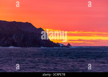Un coucher de soleil spectaculaire sur l'océan, avec une côte rocheuse silhouette sur le ciel orange et rouge vibrant. Les vagues s'écrasent contre les rochers, créant un Banque D'Images