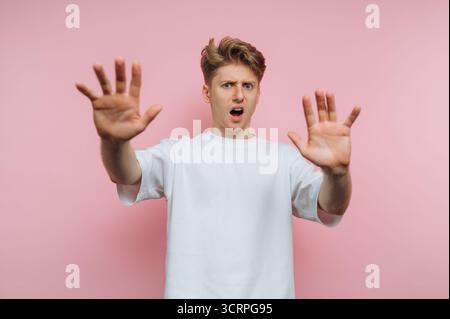 portrait du jeune homme consterné lève les mains dans le choc et l'incrédulité, capturant un moment émotionnel intense sur fond rose Banque D'Images