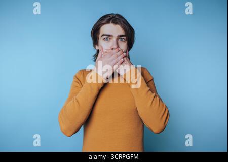 portrait d'un jeune homme choqué dans un col roulé orange couvre sa bouche avec des mains sur fond bleu Banque D'Images