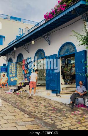 Scène de rue bleue et blanche à Sidi Bou Said, Tunisie : art mural en céramique, chemin pavé et sièges de café. Capturé le 23 septembre 2025. Banque D'Images