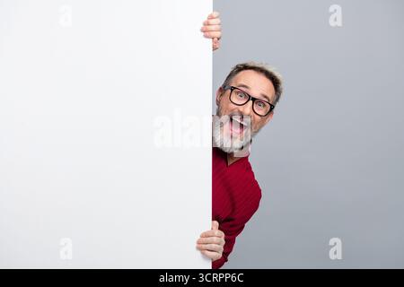 Homme d'affaires joyeux dans des lunettes jetant un œil ludique de derrière un panneau d'affichage vierge, mettant en valeur l'énergie vibrante et la joie Banque D'Images