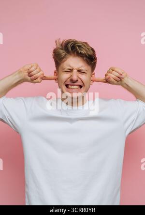 Un jeune homme aux cheveux blonds courts sourit largement en bouchant ses oreilles avec ses doigts. Il se tient sur un fond rose Uni, véhiculant un humour Banque D'Images