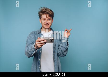 Un jeune homme se tient sur un fond bleu solide, souriant largement tout en tenant un smartphone dans une main et en faisant des gestes ludiques avec l'autre. Son c Banque D'Images