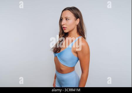 Une jeune femme portant une tenue athlétique bleue se tient confiante dans le profil sur un fond clair. Ses cheveux coulent doucement tandis qu'elle regarde avec soin Banque D'Images