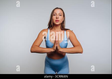 Une femme vêtue d'une tenue d'entraînement bleue pratique le yoga avec ses paumes ensemble dans un espace serein. Sa pose reflète la concentration et la tranquillité Banque D'Images