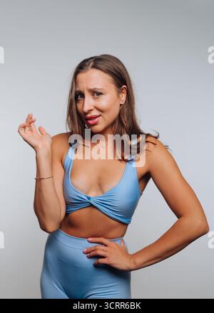 Une jeune femme portant un short top bleu et des leggings assortis prend une pose ludique dans un studio. Elle sourit, montrant sa confiance et sa personnalité Banque D'Images
