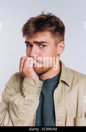 Un jeune homme semble anxieux, se mordant les ongles et paraissant inquiet. Il se tient dans un cadre intérieur simple avec un fond neutre, habillé Casua Banque D'Images