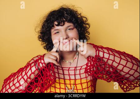 Une fille aux cheveux bouclés sourit joyeusement tout en posant sur un fond jaune vif. Elle porte un haut en filet rouge et pointe ludique sur sa joue Banque D'Images