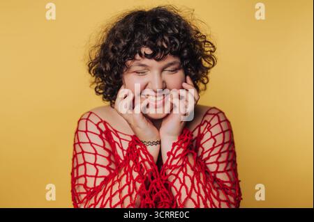 Une jeune femme aux cheveux bouclés sourit chaleureusement tout en portant une tenue en filet rouge frappante. Le fond jaune vif renforce son expression joyeuse an Banque D'Images